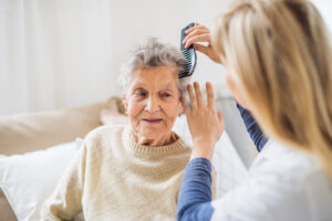 An unrecognizable healthcare professional combing the hair of an elderly woman who's family had to choose between living with family vs. assisted, sitting on a sofa at home.